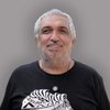 Lachender Mann mit grauen Haaren und Bart, trägt ein schwarzes T-Shirt mit gemusterter Afrika-Kontur und ein Kreuz an einer Halskette.