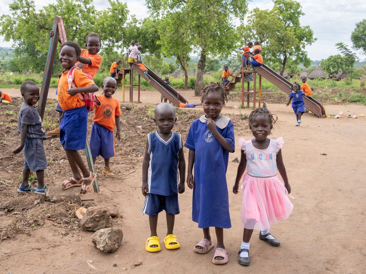 Fröhliche Kinder in einem afrikanischen Dorf. Drei lächeln im Vordergrund, andere spielen auf einer Rutsche unter Bäumen.