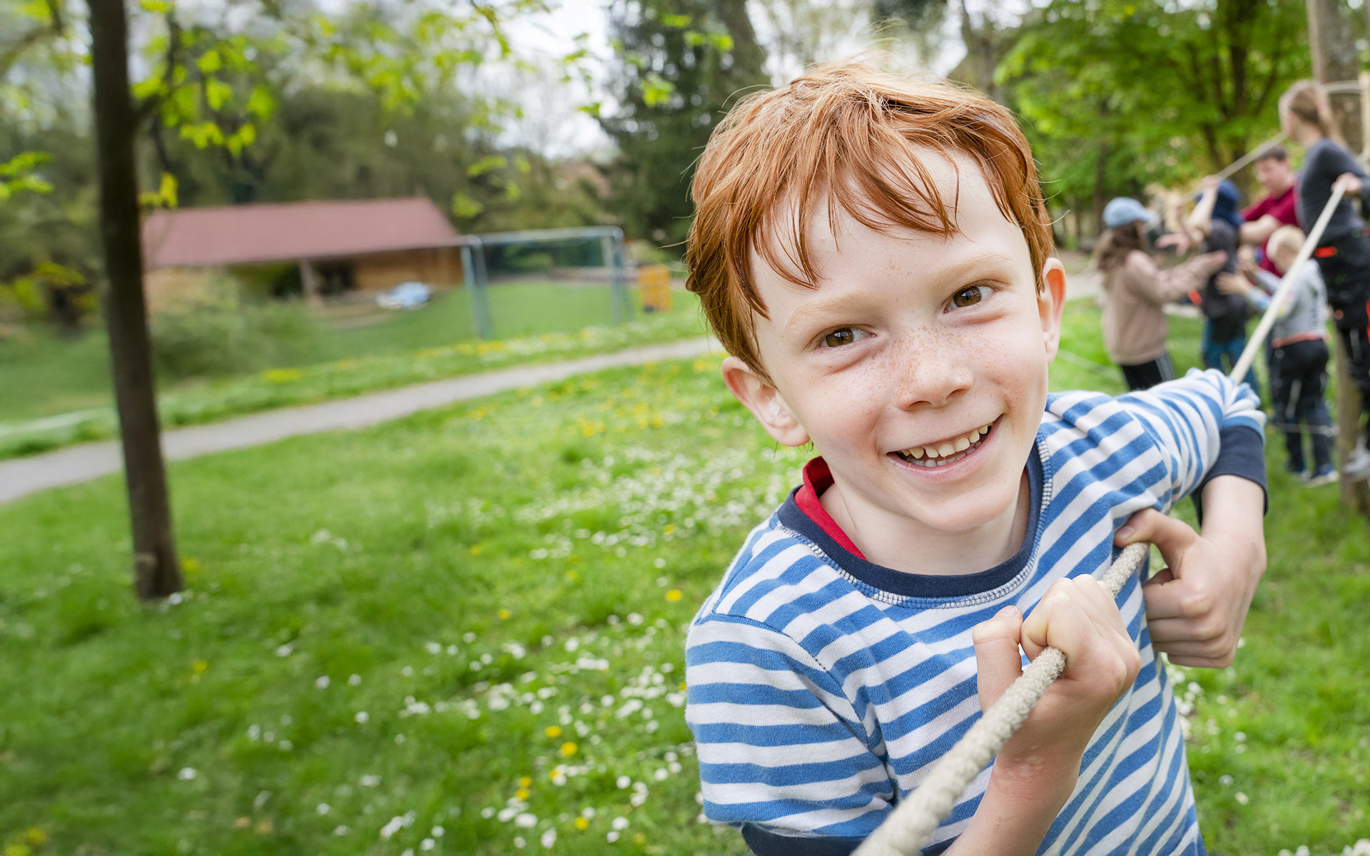 Lächelnder rothaariger Junge mit Sommersprossen hält ein Seil, spielt mit anderen Kindern bei einer Outdoor-Aktivität.