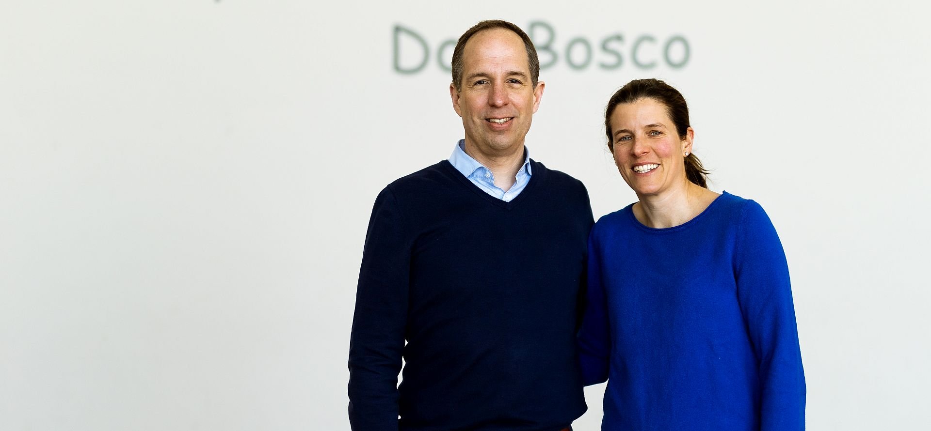 Ein lächelnder Mann im blauen Pullover und eine Frau im blauen Oberteil stehen nebeneinander vor einer weißen Wand mit 'Do Bosco'-Schriftzug.