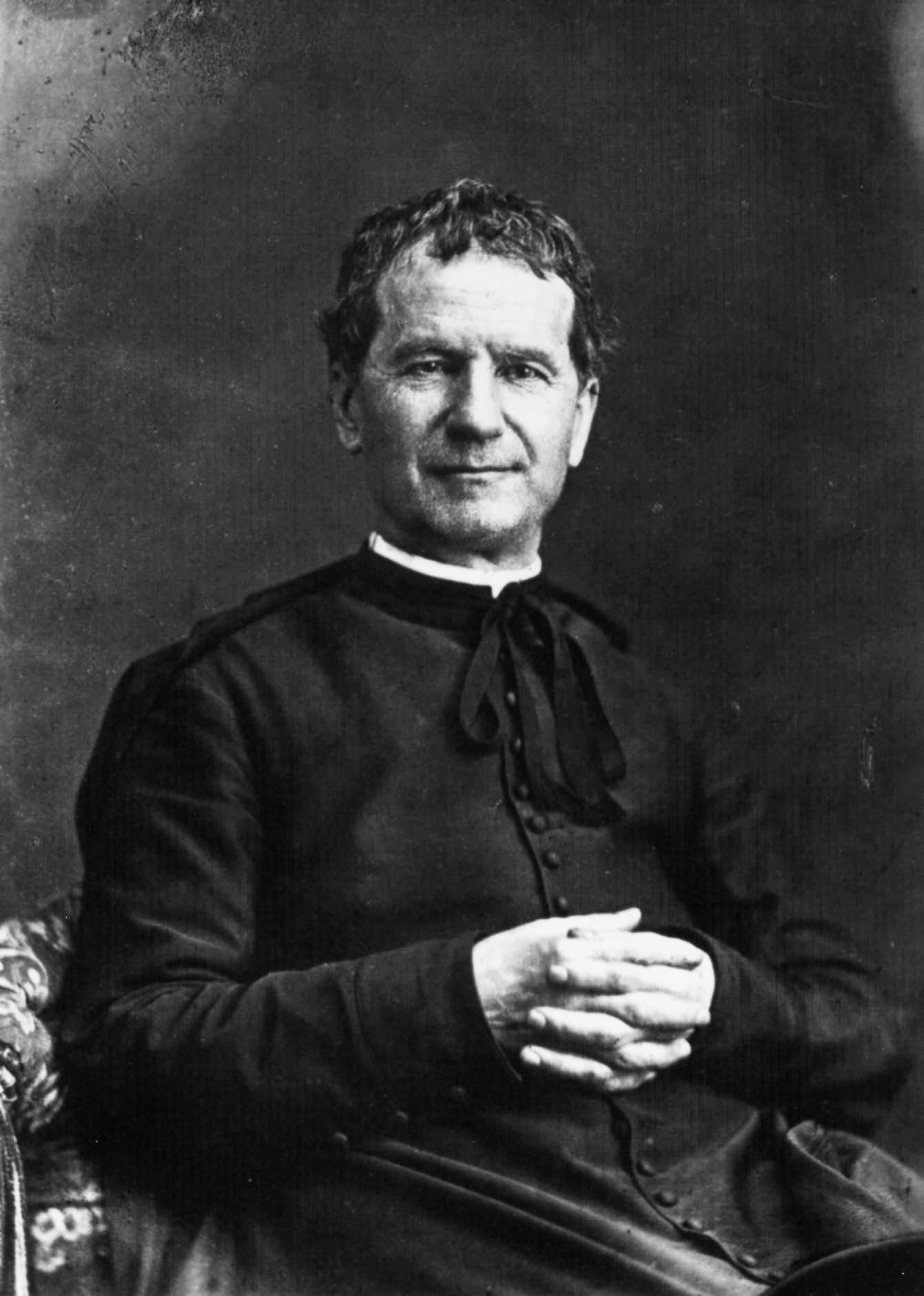 Schwarz-Weiß-Porträt des Heiligen Don Bosco, eines Priesters und Pädagogen, sitzend mit gefalteten Händen.
