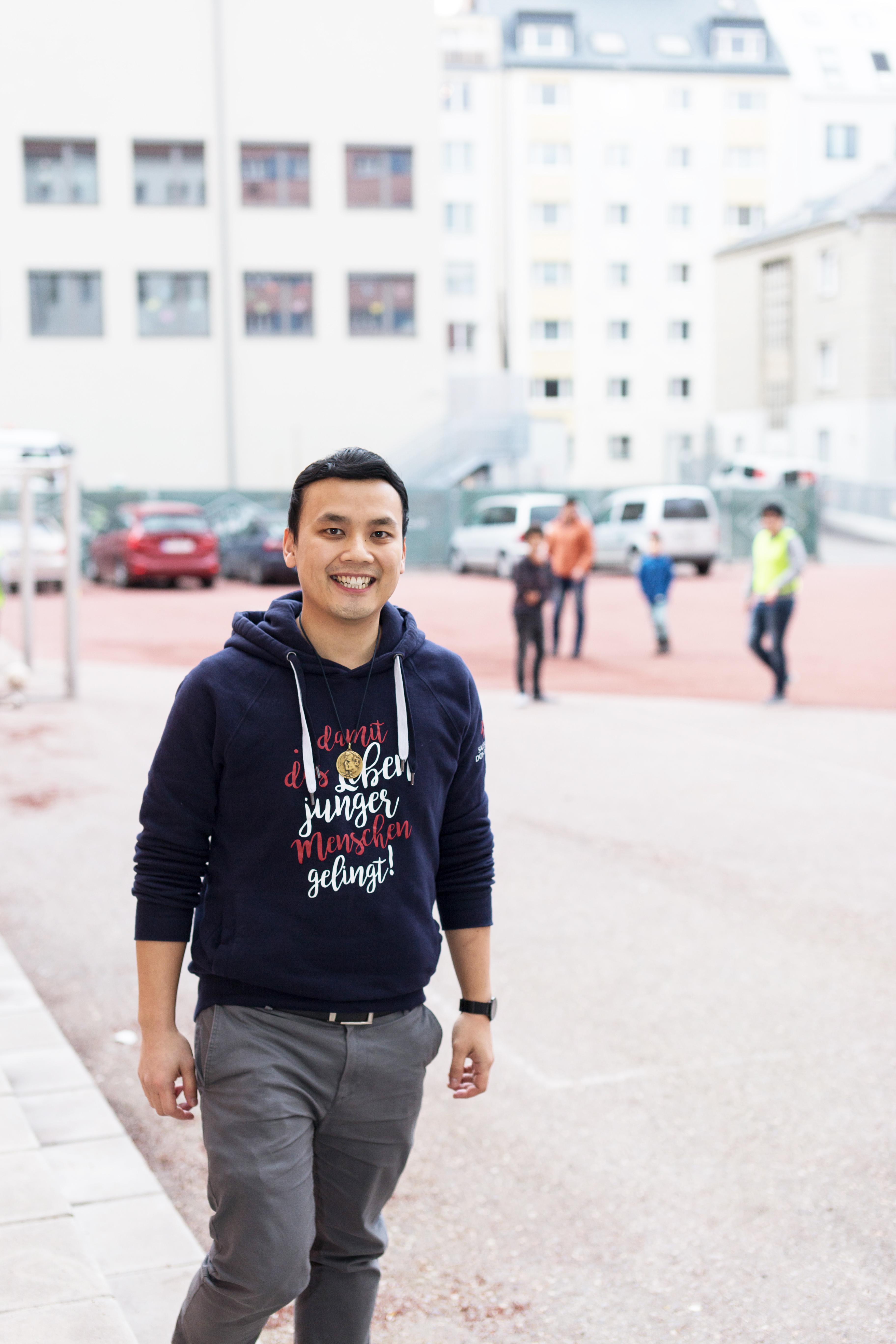 Lächelnder asiatischer Mann in blauem Hoodie mit deutschem Text 'damit das Leben junger Menschen gelingt!', im Freien.