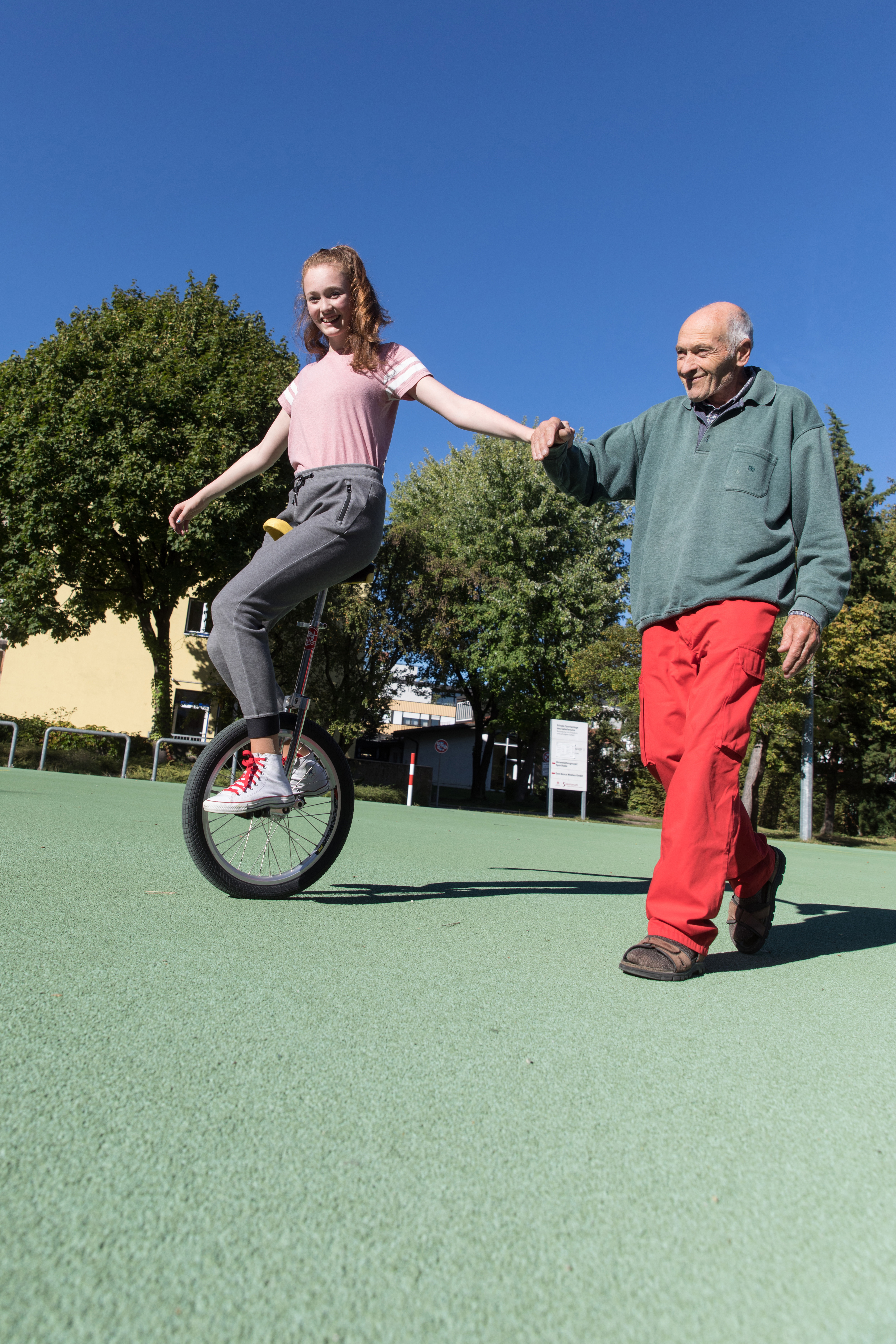 Eine junge Frau auf einem Einrad hält Händchen mit einem älteren Mann, der ihr lächelnd auf einem grünen Platz hilft.