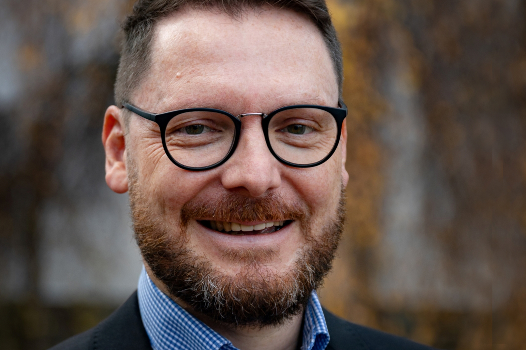 Nahaufnahme eines lächelnden Mannes mit dunklen Haaren, Bart und Brille, der ein kariertes Hemd und eine Jacke trägt.