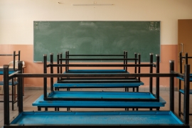 Ein Foto eines leeren Klassenzimmers. Blaue, umgekehrt aufeinander gestapelte Tische vor einer orangeroten Wand mit einer großen, grünen Tafel.
