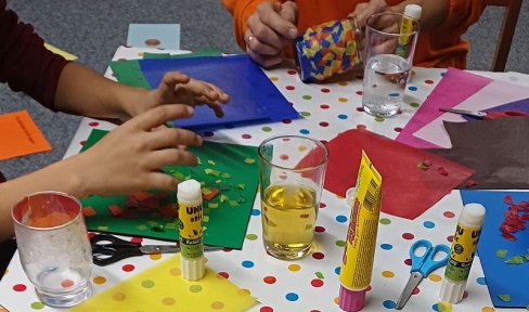 Kinder sitzen um einen Tisch und basteln Erinnerungslichter aus Gläsern und buntem Papier