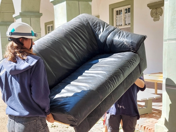Junge Freiwillige tragen ein Sofa aus den durch das Unwetter am 26. August 2023 zerstörten Unterkünften im Kloster Benediktbeuern